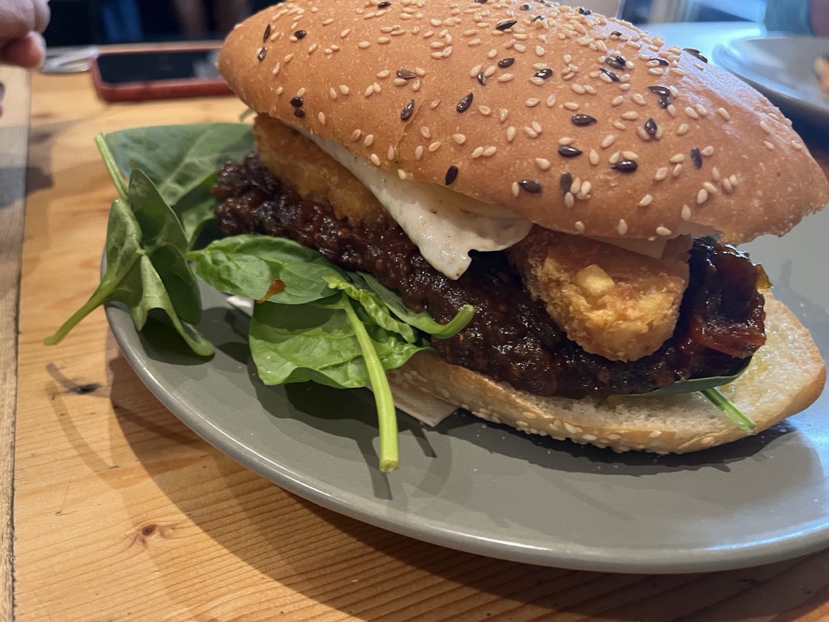 A sesame-seed bun filled with a dark patty, a round golden fritter, spinach and white sauce on a grey plate