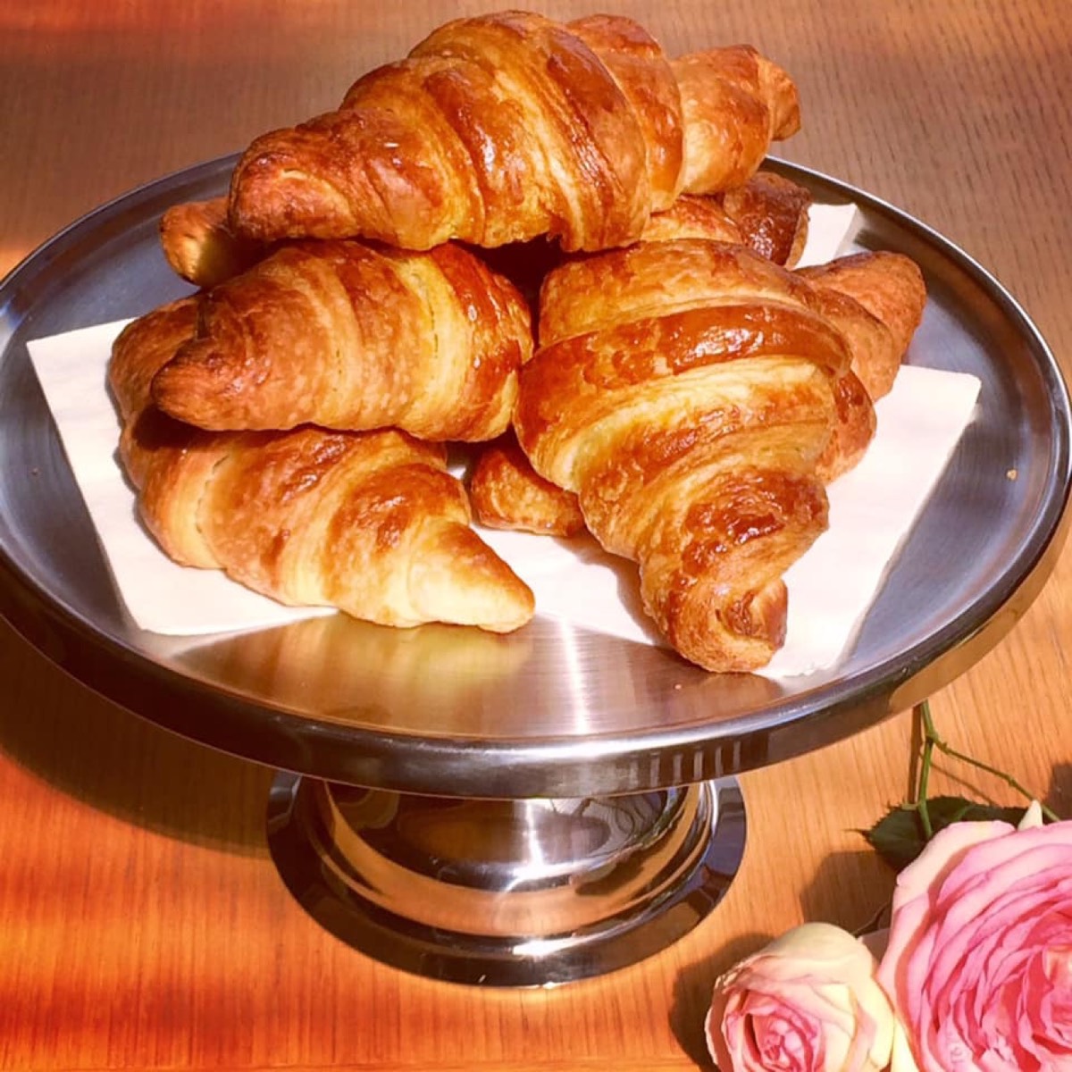 A pile of golden croissants on a steel cake stand with a pink rose beside it