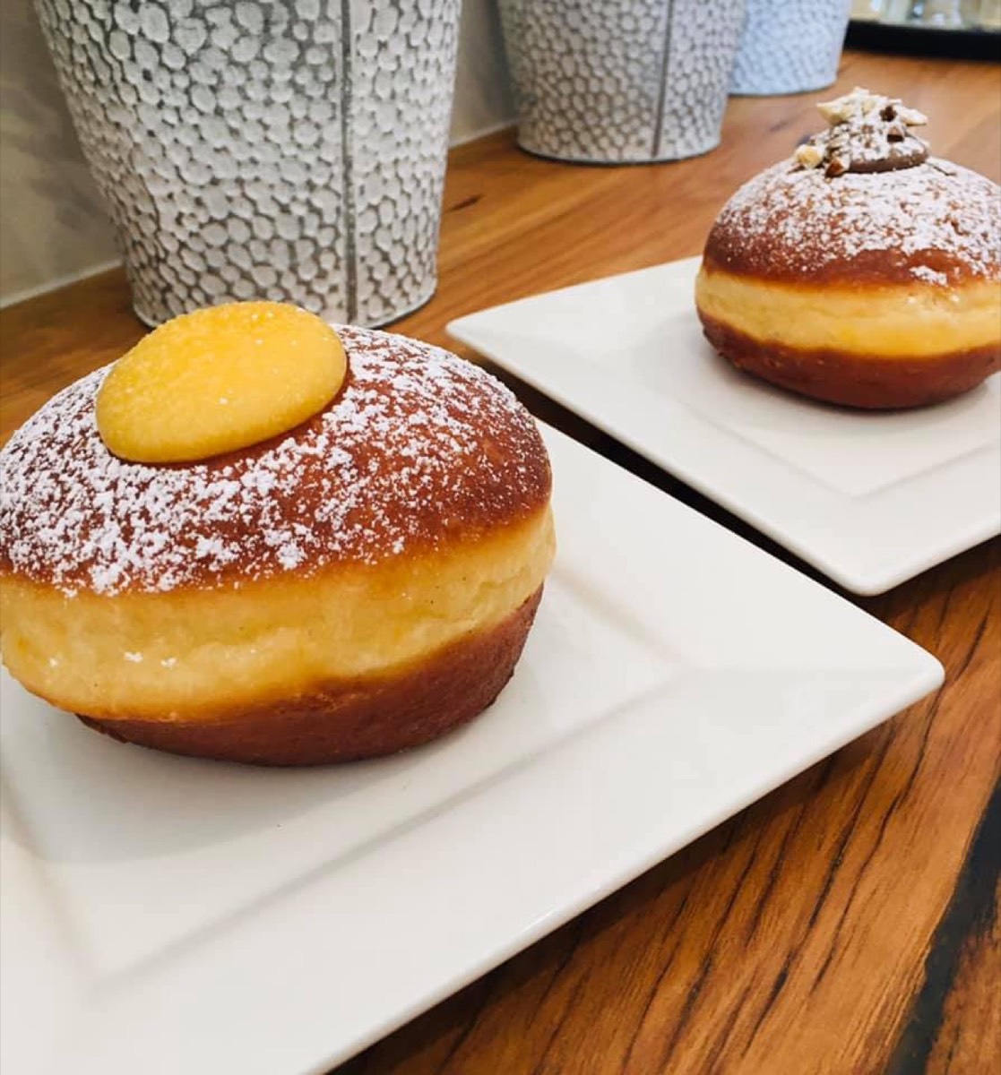 A round filled doughnut dusted with icing sugar and topped with a yellow disc, on a white square plate, with a smaller one behind