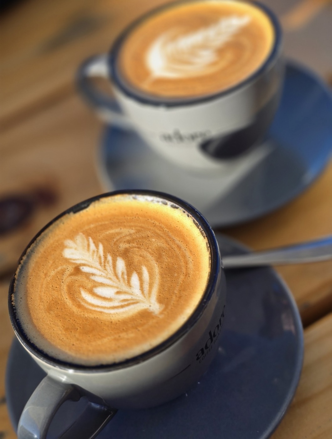 A flat white with a leaf pattern in the milk foam, in a grey cup on a saucer, with a second coffee behind it