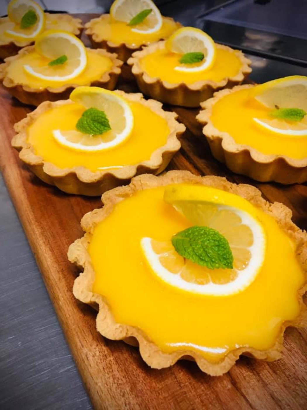 Six lemon tarts with bright yellow filling and mint leaves on a wooden board