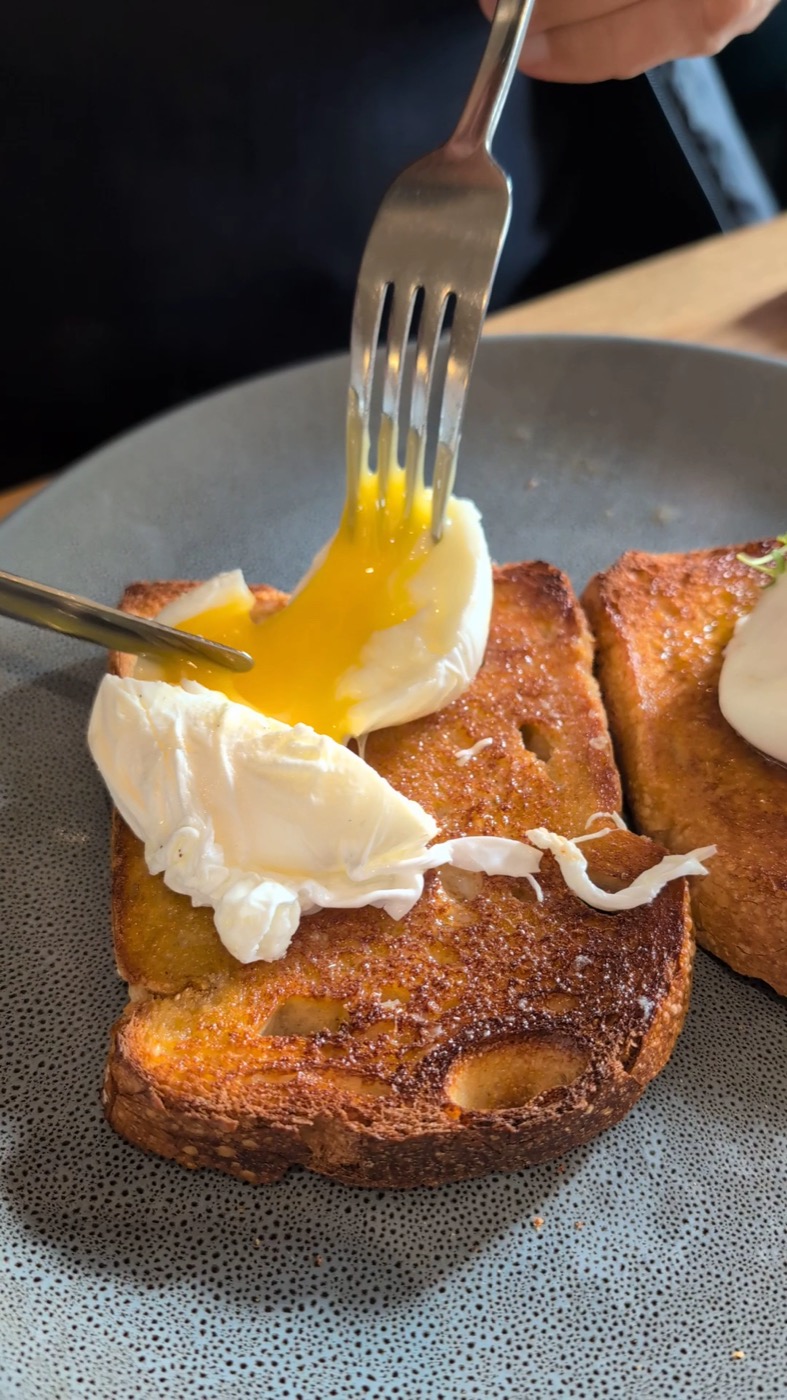 A poached egg with a runny yolk being cut with a fork, on thick slices of toast on a speckled plate
