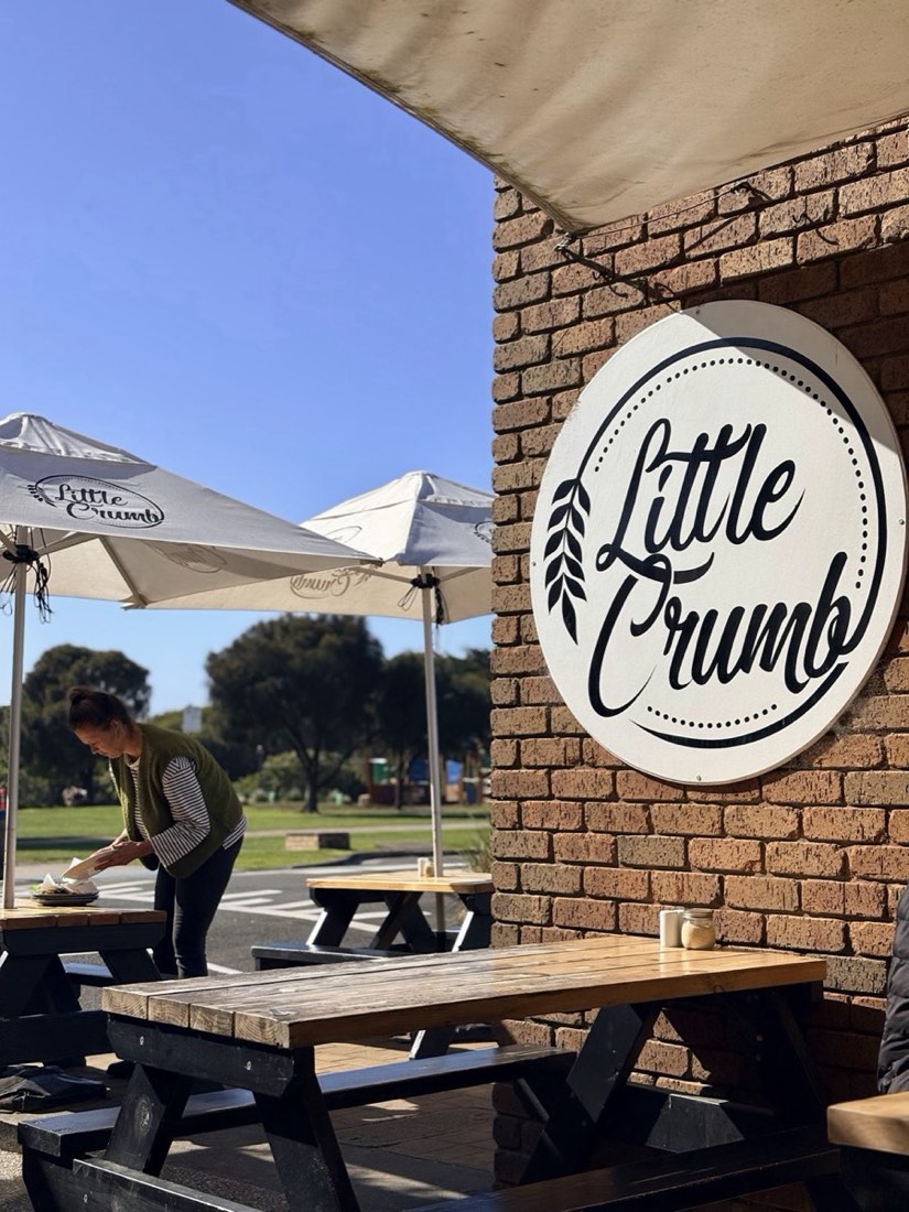 The outside of the shop: a round Little Crumb logo on a brick wall, with picnic tables and market umbrellas on the footpath