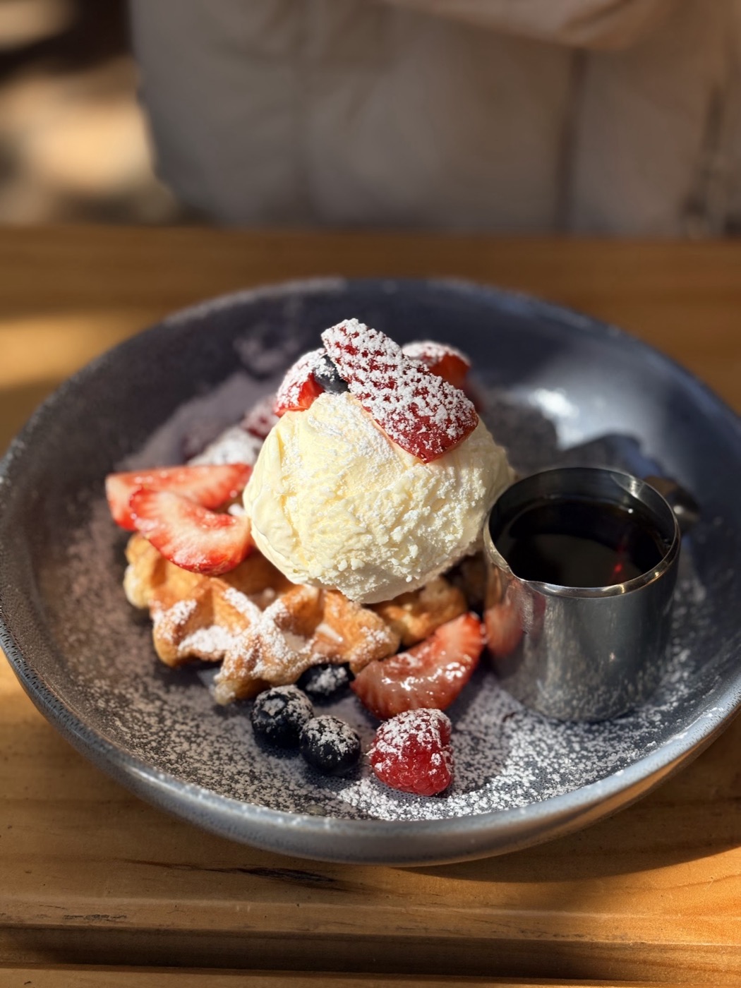 A bowl of waffles topped with a scoop of vanilla ice cream, strawberries, blueberries and icing sugar, with a small metal jug of syrup beside it